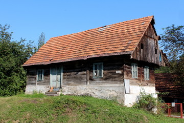 Fototapeta na wymiar Small wooden old family house with dilapidated boards and broken roof with missing roof tiles on top of small hill surrounded with grass and trees with clear blue sky in background