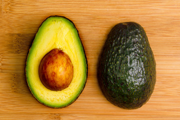 Two halves of an avocado, 1 face down and 1 face up with the pit still in place, on a bamboo cutting board.