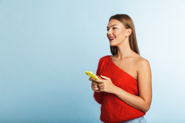 Smiling young woman posing isolated over blue wall background using mobile phone.