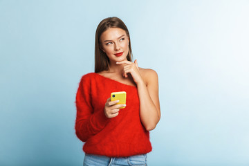 Dreaming thinking young woman posing isolated over blue wall background using mobile phone.