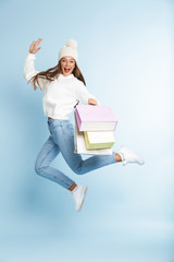 Cute young woman wearing winter hat jumping isolated over blue wall background holding holding bags.