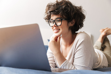 Happy young woman indoors on bed using laptop computer.