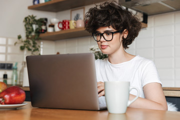Fototapeta premium Happy young woman sitting indoors at kitchen using laptop computer.