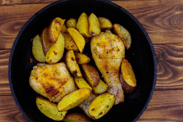 Baked chicken drumsticks with potatoes in in cast-iron frying pan on wooden table. Top view