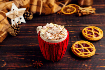 A cup of hot drink with whipped cream and powder, in a knitted cover and homemade cookies, Cezve and spices, lie on a wooden table near the checkered rug. Copy space.