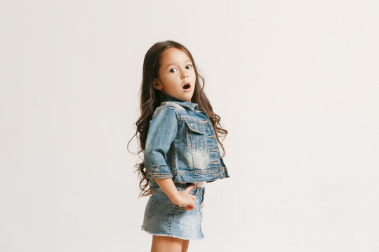 The portrait of cute little kid girl in stylish jeans clothes looking at camera and smiling, standing against white studio wall. Kids fashion concept
