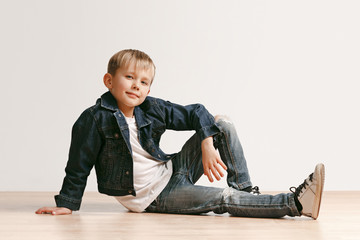 The portrait of cute little kid boy in stylish jeans clothes looking at camera against white studio wall. Kids fashion concept