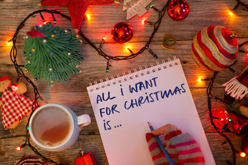 “All I want for Christmas is…” text on notebook written by young woman with colorful gloves in festive decor 