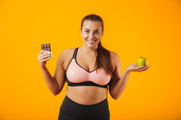 Image of happy overweight woman in tracksuit holding chocolate bar and apple in both hands,...