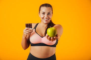 Image of sporty chubby woman in tracksuit holding chocolate bar and apple in both hands, isolated over yellow background