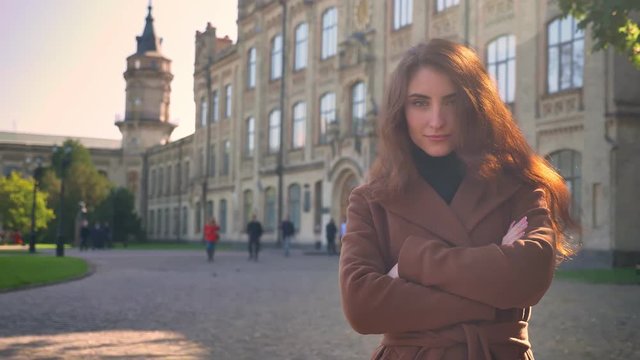 Amazing caucasian brunette girl is standing outdoors straight and looking at camera with natural relaxed position, buildings on background