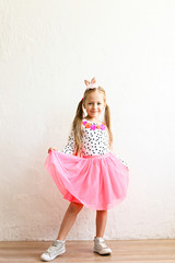 Portrait of cute little five year old girl with long blond hair wearing easter bunny ears on head, colorful polka dot shirt and pink tutu skirt. White wall backgroung, copy space, close up.