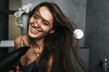 Photo of joyful woman with long dark hair and healthy skin drying her hair, while standing in bathroom