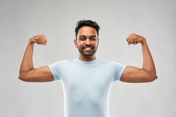 power, strength, sport and people concept - happy young indian man showing biceps over grey...