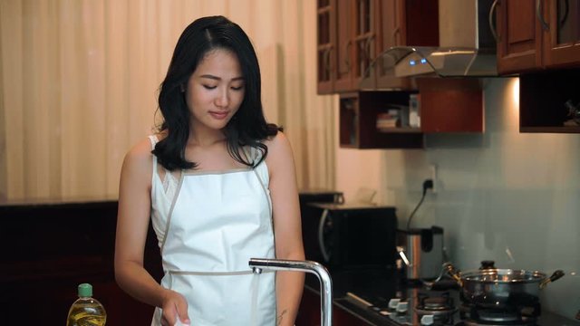 Young attractive Asian housewife standing in home kitchen near sink, washing dishes with detergent and smiling cheerfully 