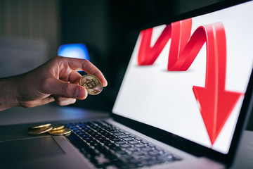 Side view of hand holding bit coin. On computer screen red arrow pointing down.