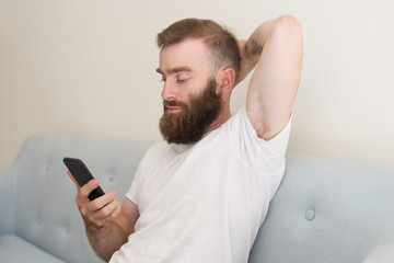 Focused man browsing on smartphone and sitting on sofa. Handsome guy keeping hand behind head and relaxing. Communication concept. Front view.