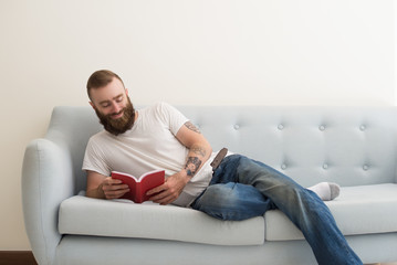 Smiling bearded man lying on sofa and reading book. Handsome guy having rest. Leisure and literature concept. Front view.