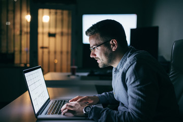 Caucasian focused employee typing on laptop while sitting in the office late at night.