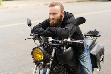 Fototapeta na wymiar Portrait of man sitting on motorbike with pensive expression. Young Caucasian biker resting on his bike outdoors. Transportation concept