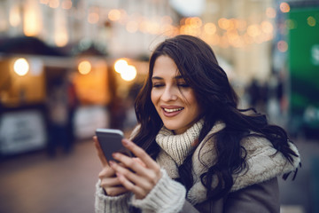 Close up of young smiling beautiful Caucasian woman using smart phone on the street on cold weather.