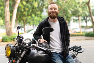 Fototapeta na wymiar Portrait of smiling young man sitting on his motorcycle. Happy biker posing with motorbike outdoors. Biker lifestyle concept
