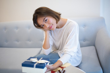 Content Asian woman in casual sweater holding Christmas gift and looking at camera. Calm girl sitting on comfortable sofa in living room. Celebration concept