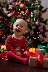 Happy baby in red pajamas on Christmas morning unpacks presents in front of the Christmas tree on a dark wooden floor