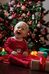 Happy baby in red pajamas on Christmas morning unpacks presents in front of the Christmas tree on a dark wooden floor