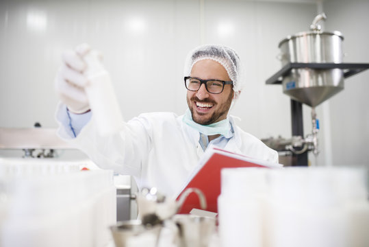 Smiling scientist holding bottle with creme in one hand and test results in other hand while sitting in the lab.