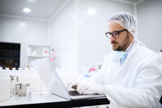Scientist using laptop for entering test results while sitting in the lab. On desk cosmetic products.