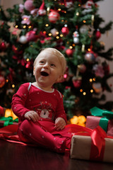 Fototapeta na wymiar Happy baby in red pajamas on Christmas morning unpacks presents in front of the Christmas tree on a dark wooden floor