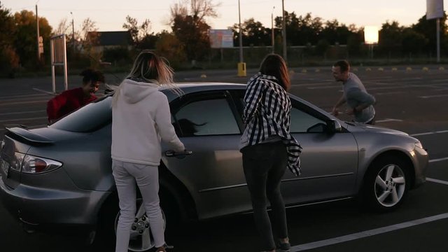 Rare view of a multirace trendy young four friends are running by mall parking zone and sit into the silver coloured car. Two couple having time together. Evening city dusk