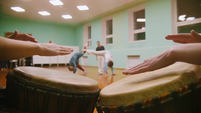 Playing a musical instrument jembe or atabaque on background male fighter performs martial tricks with dance elements in the sport gym