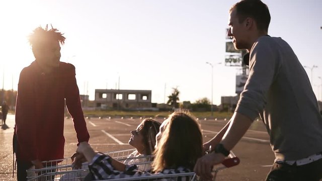 Young friends having fun on a shopping carts. Multiethnic young people playing with shopping cart. Boys standing near the shopping corts with their girlfriends. Outdoors. Sun shines