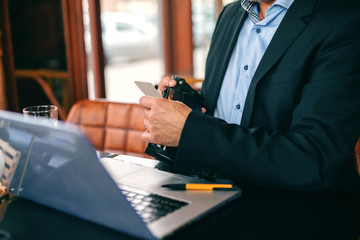 Close up of senior businessman taking credit cart out of wallet while sitting in cafeteria. On the desk laptop and glass of water.