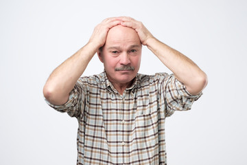 Miserable and gloomy man, frowning and looking unsatisfied while holding head, standing over gray background. He is puzzled by the big problems that have come over him.