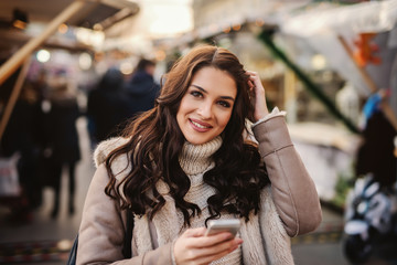 Beautiful Caucasian woman with long brown hair standing on the street on cold weather, holding smart phone and looking at camera.
