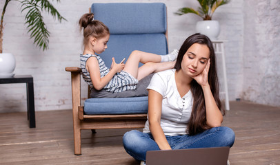 Mother working with laptop and cute little girl laying on the armchair and playing games on the smartphone