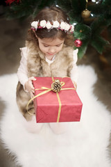 A pretty little girl kid holds a big Christmas gift in front of a Christmas tree. Christmas...