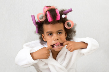Black girl with curlers with hairpins in her hair. A little girl in a white coat spun pink curlers...