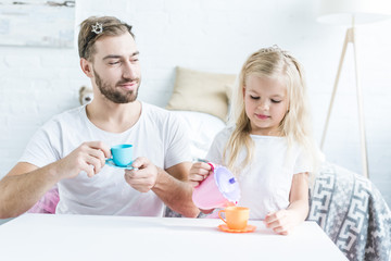 smiling father holding toy cup and looking at little daughter pouring tea from toy kettle