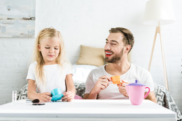 happy father with makeup and cute little daughter smiling each other while playing with toy dishes at home
