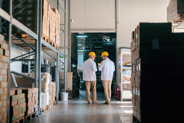 Two workers walking in the storage. Boxes on shelves. Back turned.