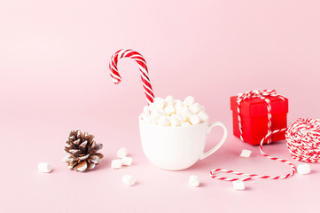 White mug with marshmallows Candy Cane, red gift box, pine cone, decorative lace on pink background...