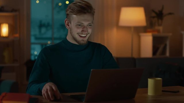 Handsome Smiling Man Works on a Laptop while Sitting at His Desk at Home. Portrait of a Young Freelancer Works on Computer in His Cozy Living Room with Warm Evening Lighting in Background.