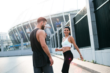 Running with cheerful smile. Rear view of young beautiful couple in sportswear looking at each other while standing against industrial city view