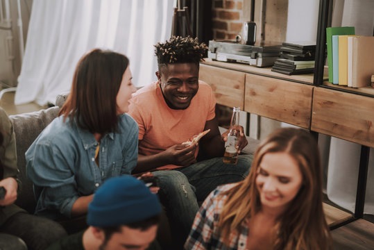 Having fun with friends. Cheerful afro american guy holding bottle of beer and slice of pizza while sitting on couch with smiling girl