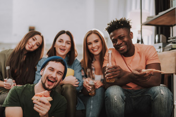 Portrait of smiling friends sitting on couch and holding bottles of beer. Focus on bearded man in hat with slice of pizza