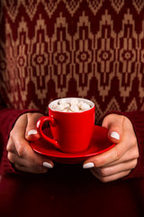 Hot chocolate with marshmallows in a red cup in the hands of a woman with a white manicure in a bardish sweater. Winter drink concept